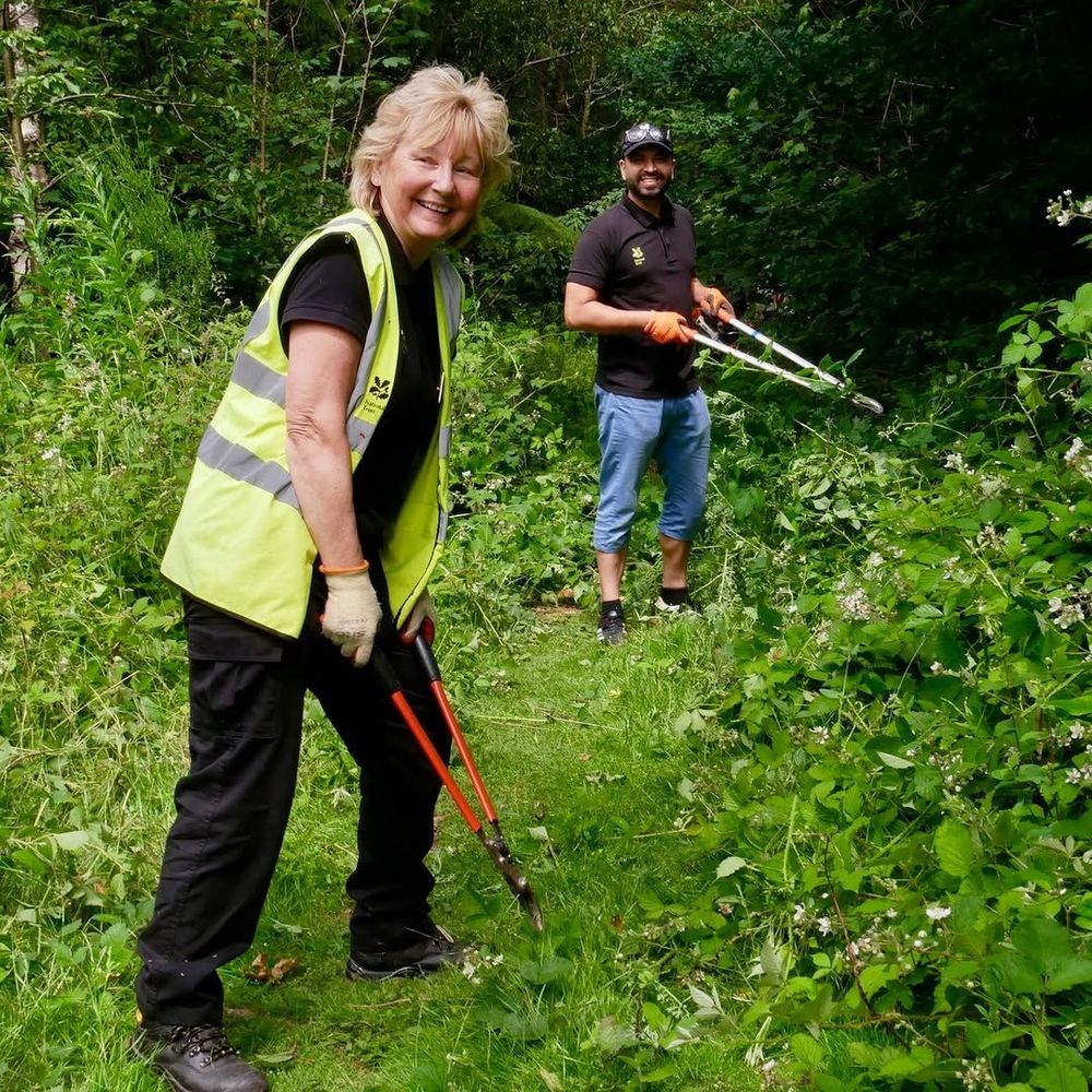 A woman in a high-vis jacket with snippers and a man with snippers in a cap behind her. They're standing amongst wild undergrowth