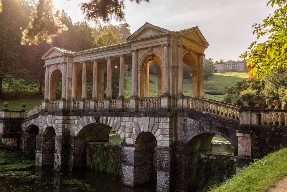 The Palladian Bridge at Prior Park at golden hour, its grand stone columns lit in the evening sun over dark water beneath, with the foliage of green trees framing it in the sunshine. 