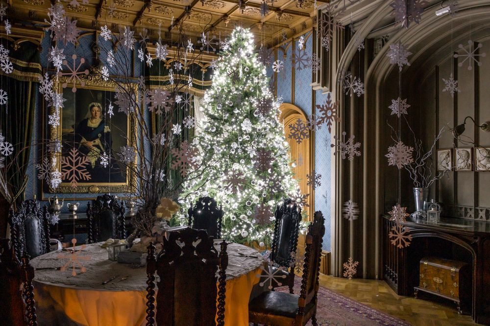 A tall Christmas tree, lit up with white lights in a room with a dining table and chairs. Snowflake decorations hang down from the ceiling