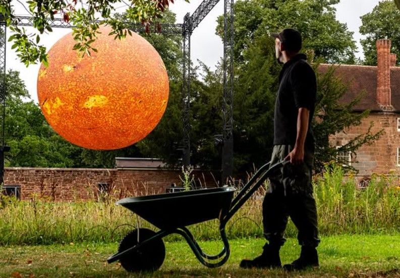 A gardener pushing a wheelbarrow pauses to look across the walled garden towards a large sun.