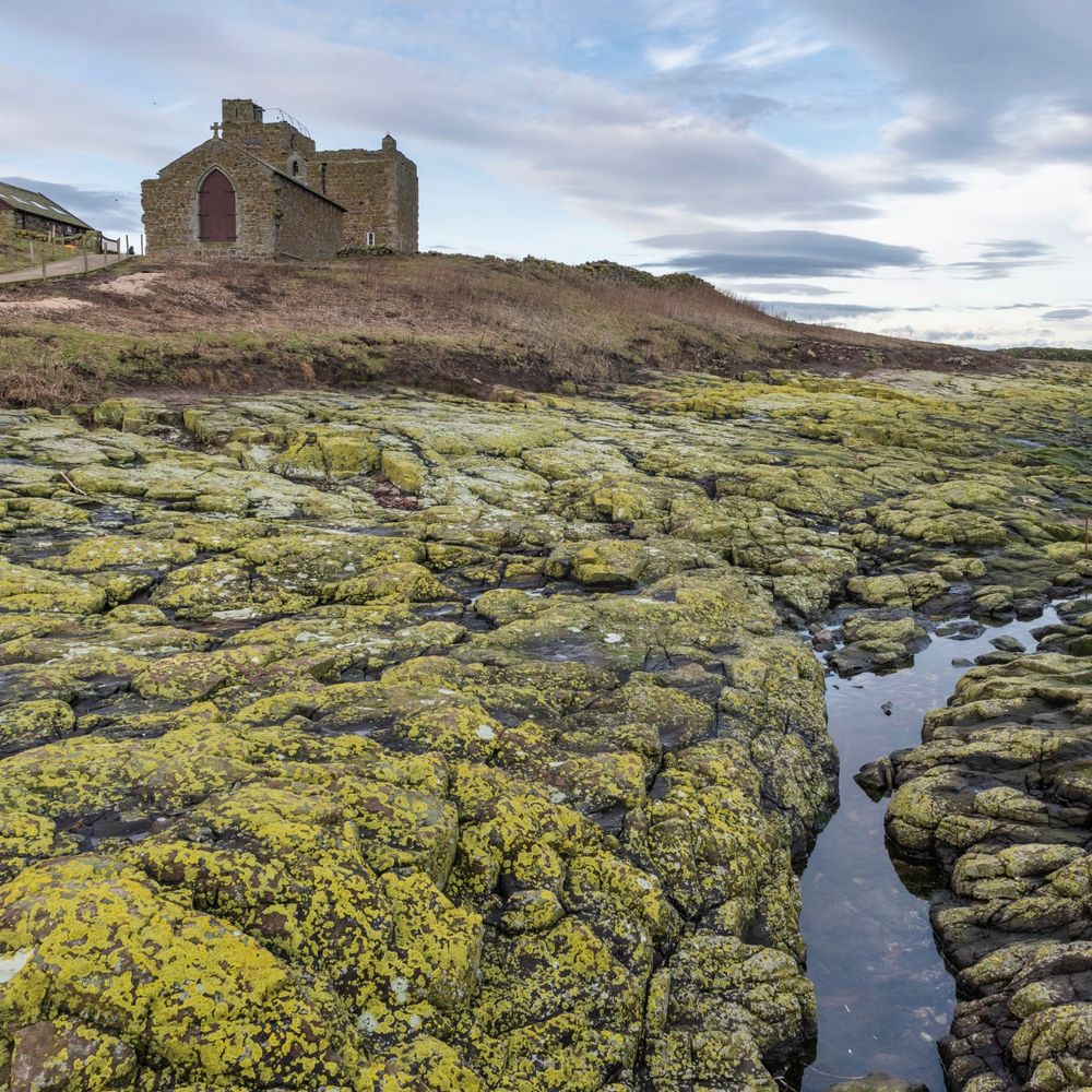 An image of the Farne Islands. A rocky coastline, punctuated by sea water, is in the foreground. An old, stone building stands at the top on the small hillside