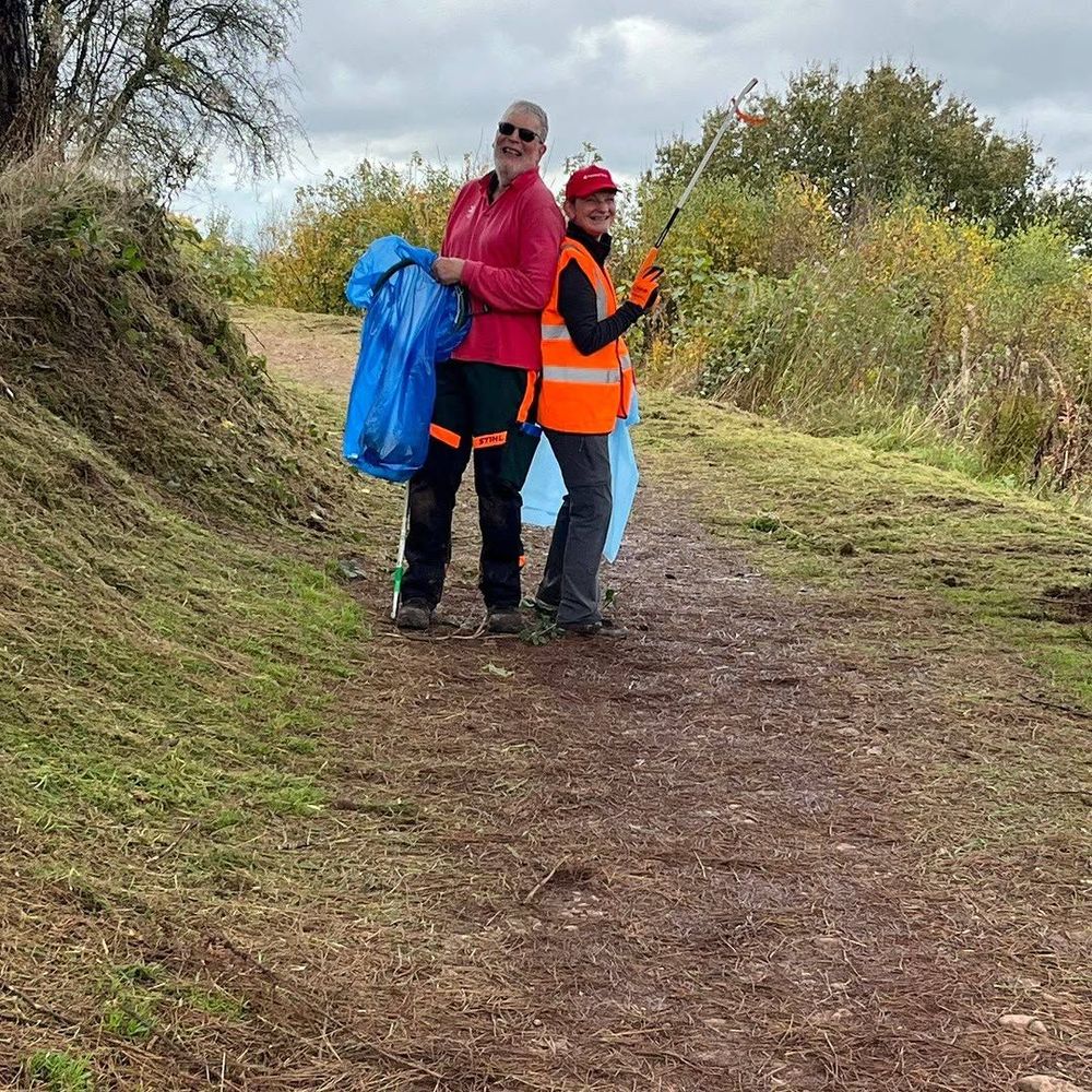 A man and a woman pose back-to-back, holding litter pickers and plastic bags. They stand on a grassy shrub-lined path