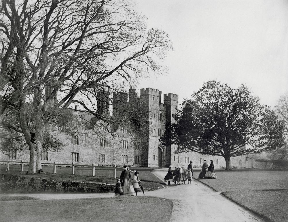 A black and white historic photo showing Victorian-era visitors to Knole in Kent. The visitors are walking down the pathways leading to the front of the historic house.