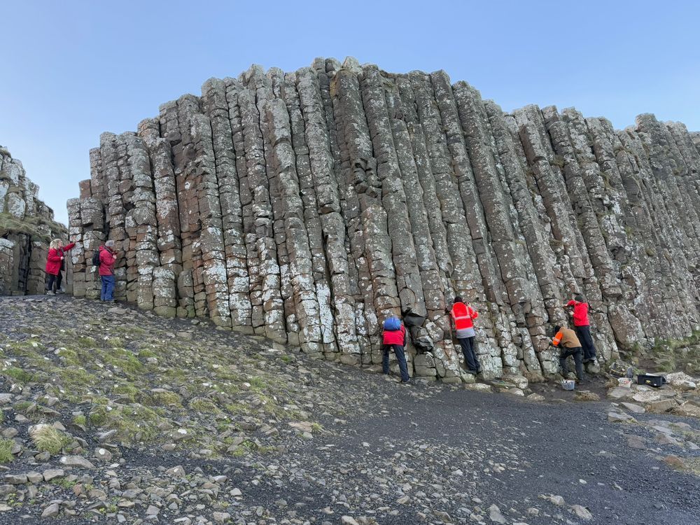 A section of the Giant's Causeway rocks. A group of people are at the base, facing towards the rocks. Most wear bright red/orange jackets. All are busy at work.