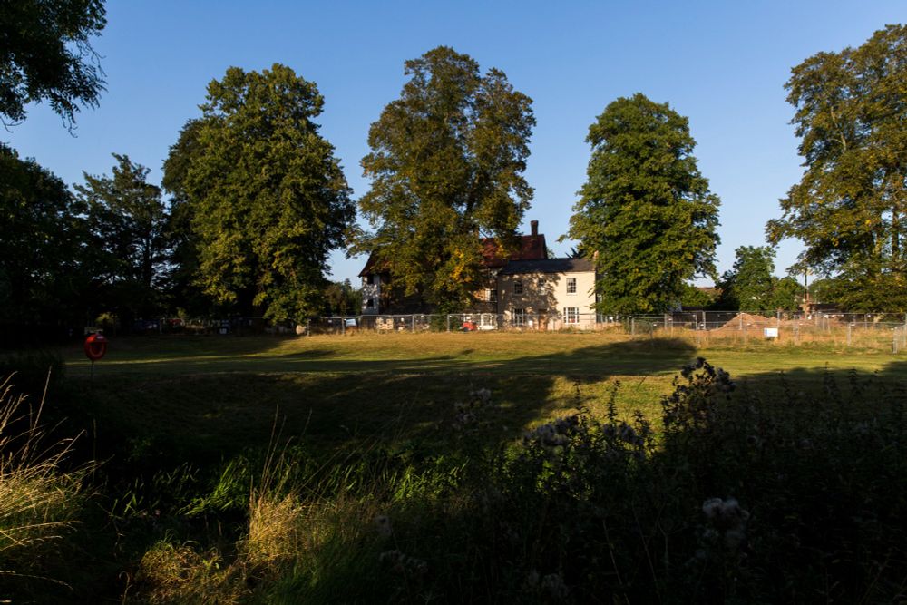 On a sunny day, green grass and green, tall trees frame the image alongside some blue sky. A large home can be seen in the distance
