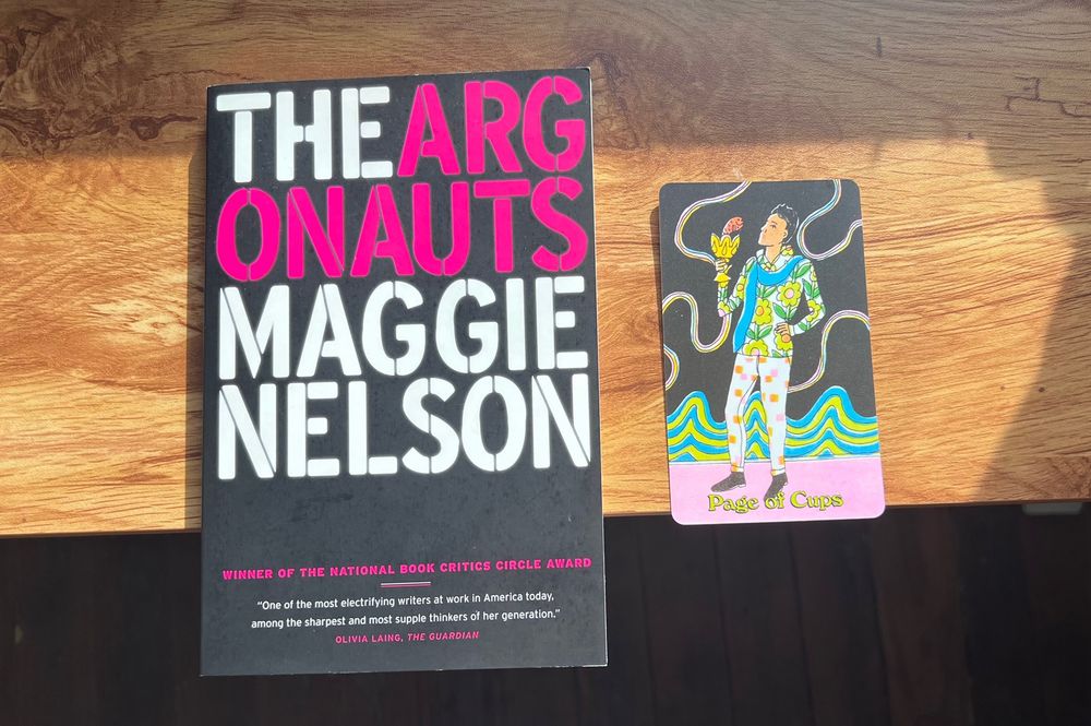 A copy of the book The Argonauts on a sunlit desk beside a page of cups tarot card featuring lots of bold colors and prints 