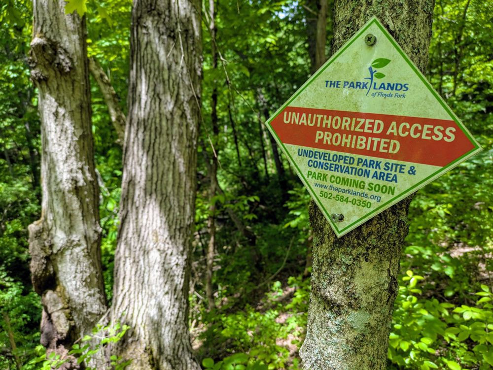 A white metal diamond sign screwed into a tree in the middle of a forest. It says "unauthorized access prohibited" in white text on red background, with some details about an upcoming park and conservation area below 