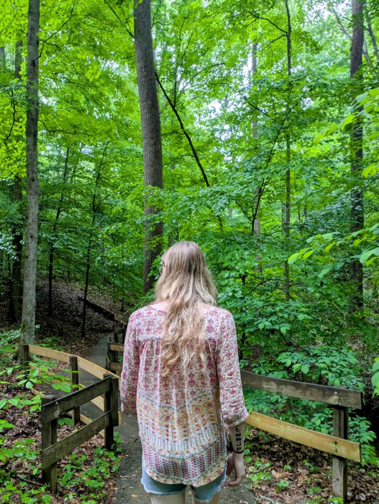 Xia is wearing a light and colorfully patterned top with denim shorts. She is walking down a small paved path in the forest lined by timber guardrails. There is a lot of greenery all around her, glistening from a recent rain fall.