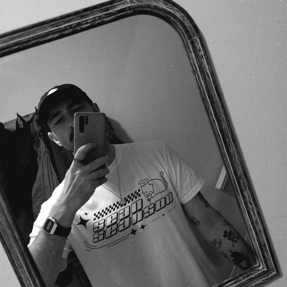 Guy taking photo of himself in a mirror wearing a white shirt that says sean seanson it has a cat on it