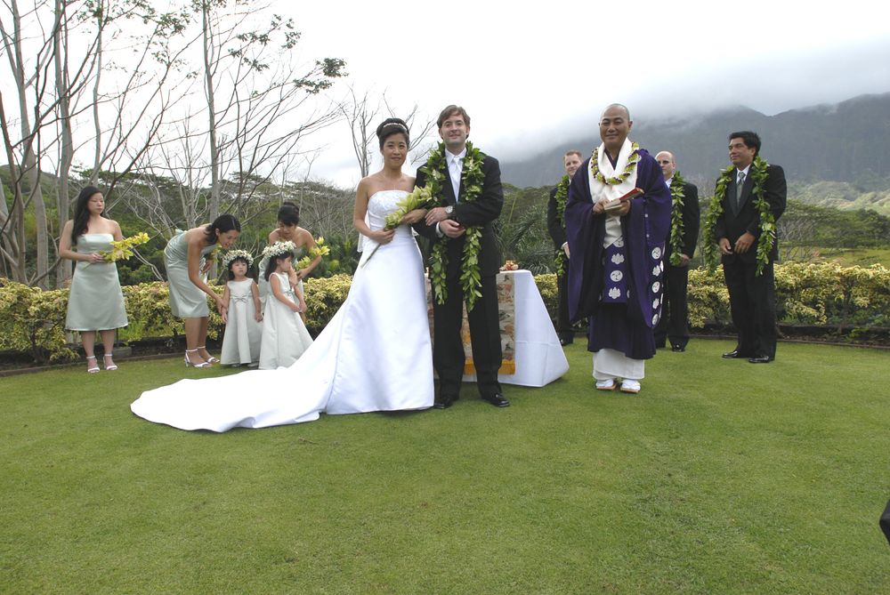 David and Sue stand on a grassy patch in wedding attire with the Koolau mountains in the background. Next to them is Bishop Dean Zenei Okimura in a purple robe. Behind them are bridesmaids, groomsmen and two flower girls.
