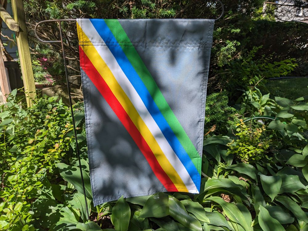 A Disability Pride garden flag hangs vertically from a thin black metal frame in the dappled sunlight, surrounded by greenery. The flag has red, yellow, white, blue, and green stripes which cut diagonally top left to bottom right across the center of a charcoal grey background.