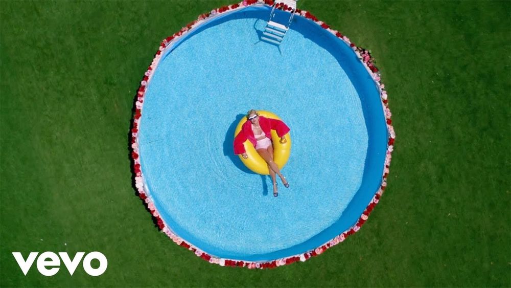 Taylor in the YNTCD video, shot from above she is wearing a pink bikini and a furry pink coat, resting on a yellow pool floating in the middle of a blue pool ringed with flowers.
