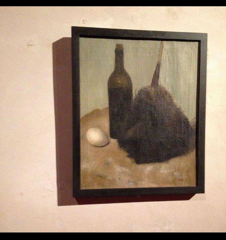 Auf einer beigen Wand hängt ein Gemälde in schwarzem Rahmen. Auf dem Gemälde ist links ein weißes Ei,daneben eine gründliche Flasche und rechts ein Staubwedel zu sehen.