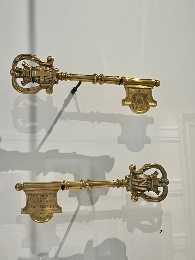 Deux clefs en bronze doré accrochées à un mur blanc, l’une avec les armoiries de la maison de Savoie et l’autre, avec le N et la couronne de Napoléon III, commémorant l’annexion de la Savoie et de son ancienne capitale à la France.
Dépôt du musée des beaux-arts de Chambéry.