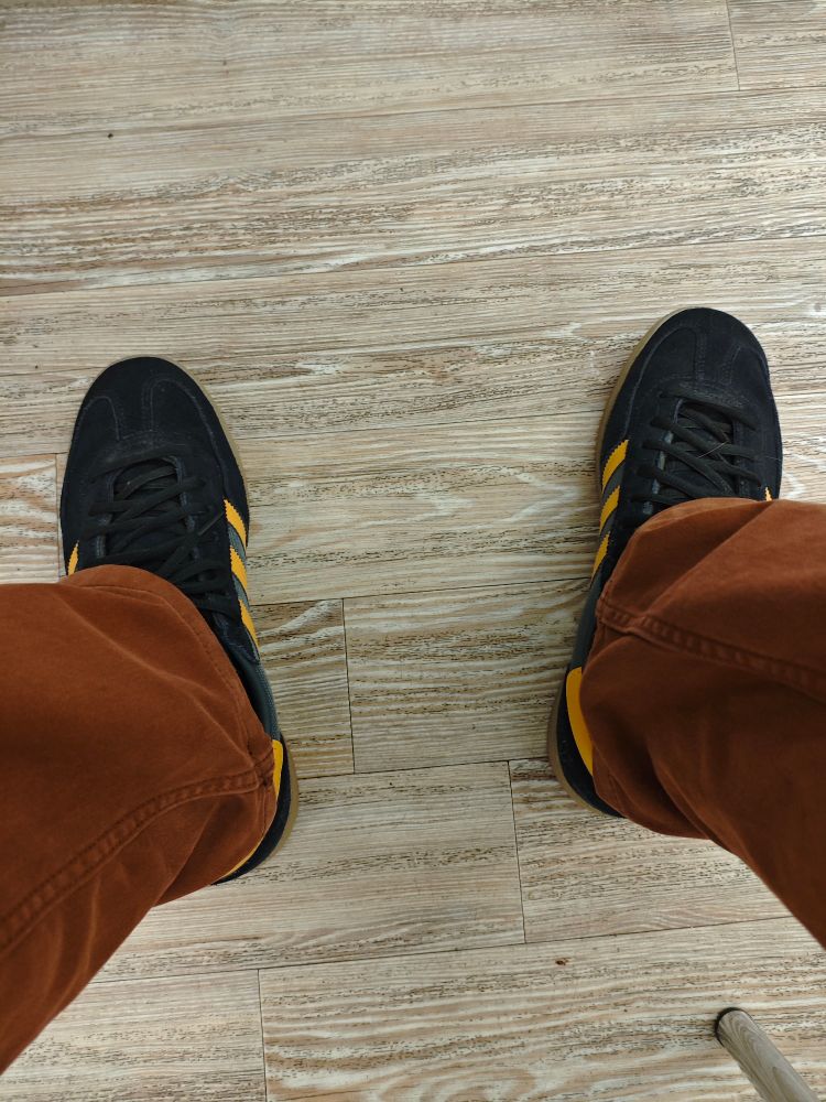 Mittelbrauner Holzfußboden, darauf Füße in schwarzen Adidas-Sneakern mit gelben Streifen und rostbrauner Hose.