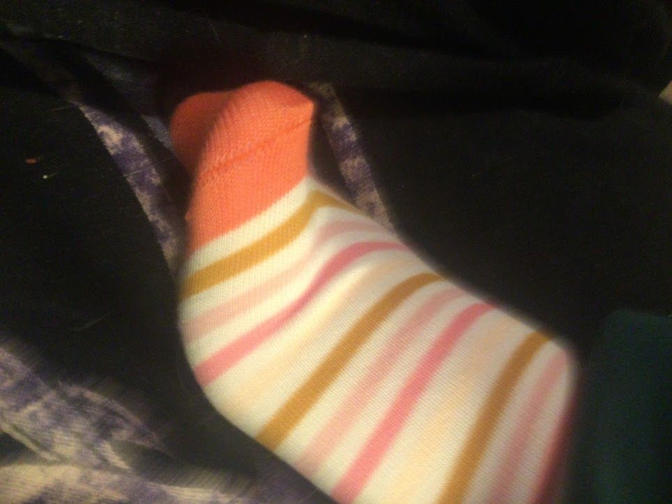 An arched foot, the ball of the foot stretched at an angle away from the camera, wearing a tight, practically sheer woman's sock, with colorful stripes. Pink, beige, mustard yellow, cream, salmon and a bright orange are the sock's colors.