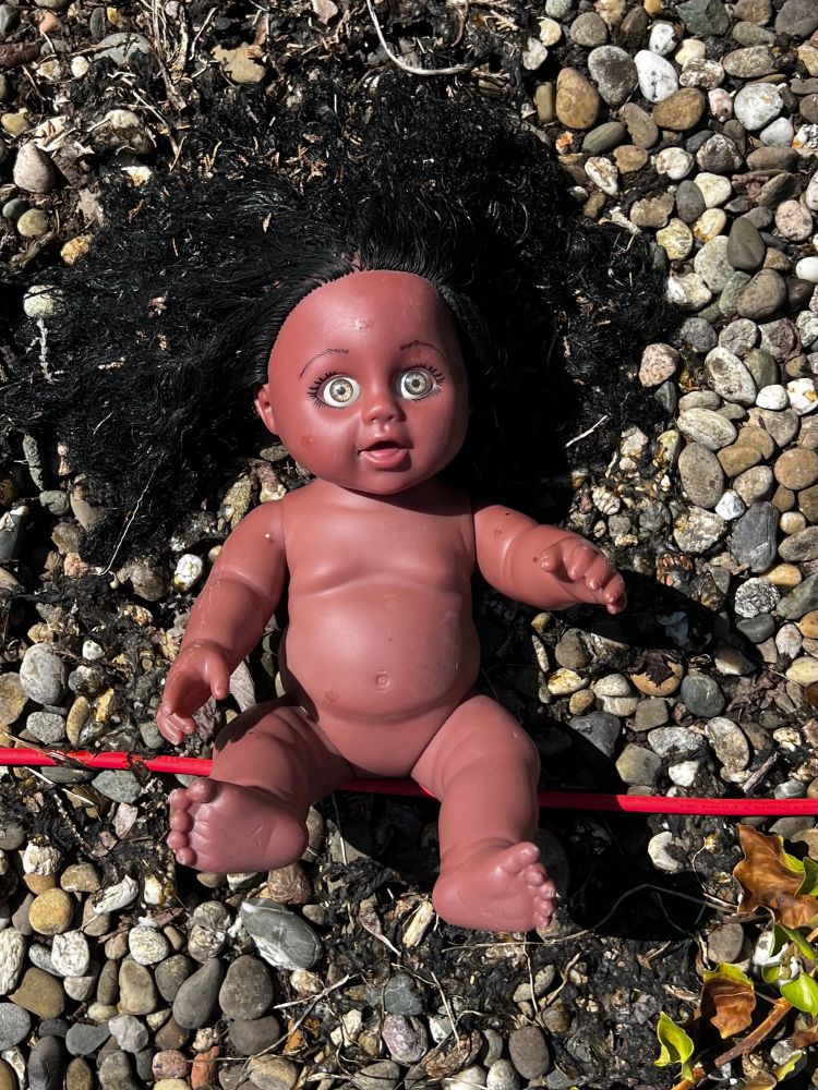 Nahaufnahme. Eine dunkelhäutige nackte Baby Puppe mit schwarzen Haaren und weit aufgerissenen, irre aussehenden Augen liegt auf einem mit Steinen belegten Garagendach.