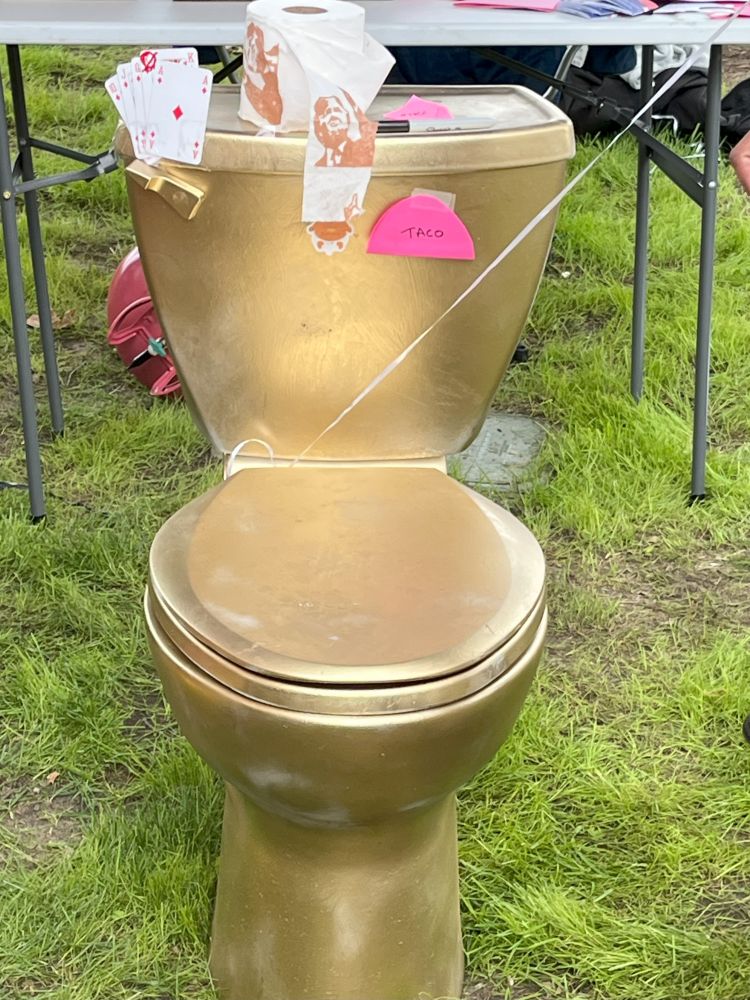 Image of a gold painted toilet. On top of it is a royal flush with the king crossed out, a pink taco stuck to it, and a roll of toilet paper with brown images of Trump.