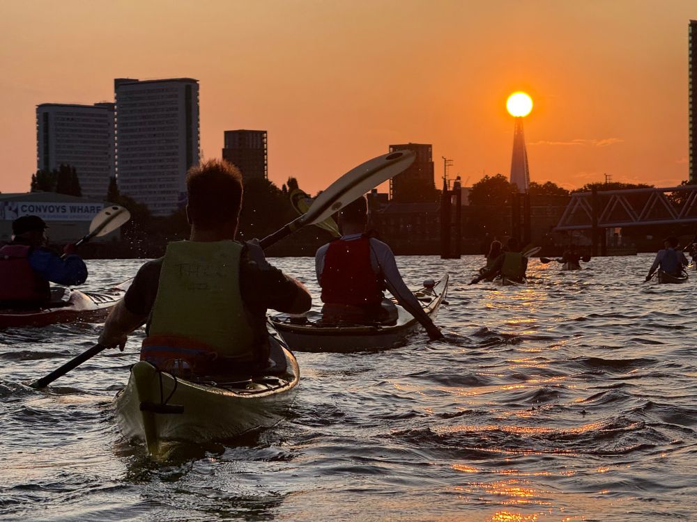 the sun is a super warm orange and just at the very top of the shard skyscraper. In the foreground there’s kayakers paddling in the Thames