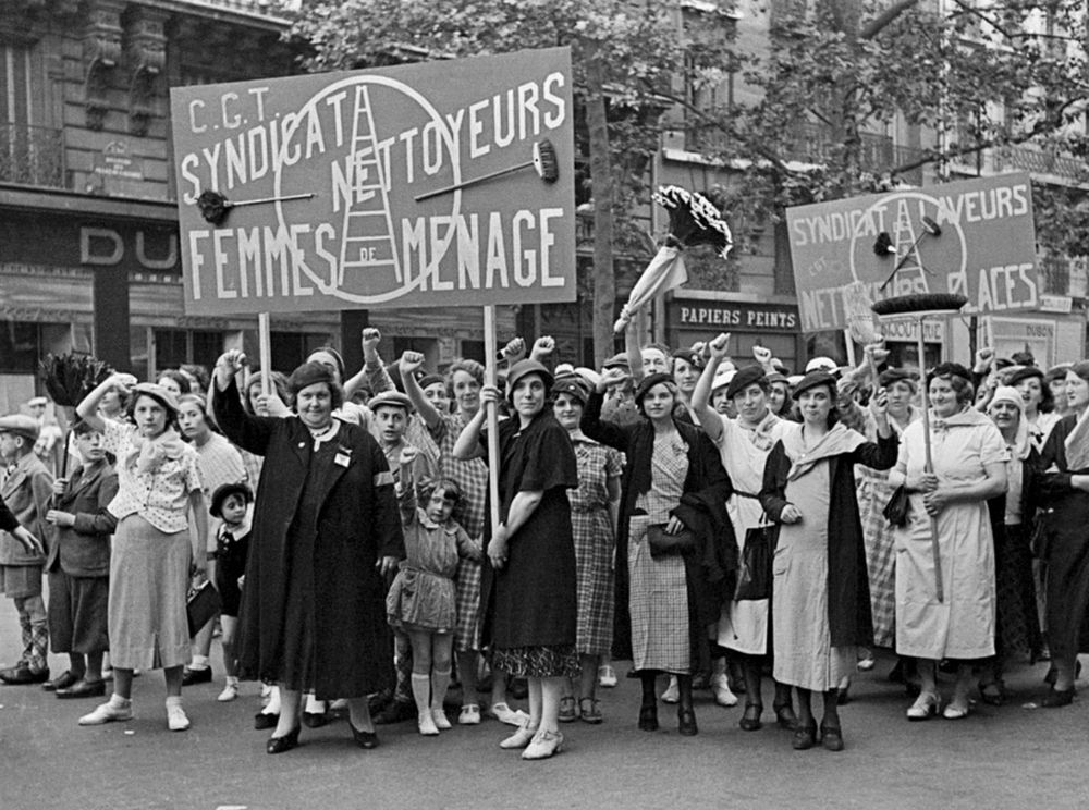 Photo du 14 juillet 1936 sous le Front populaire. Des femmes vêtues de tabliers et portant des estensiles de travail, femmes de tous âges, regardent l'objectif et lèvent le poing.