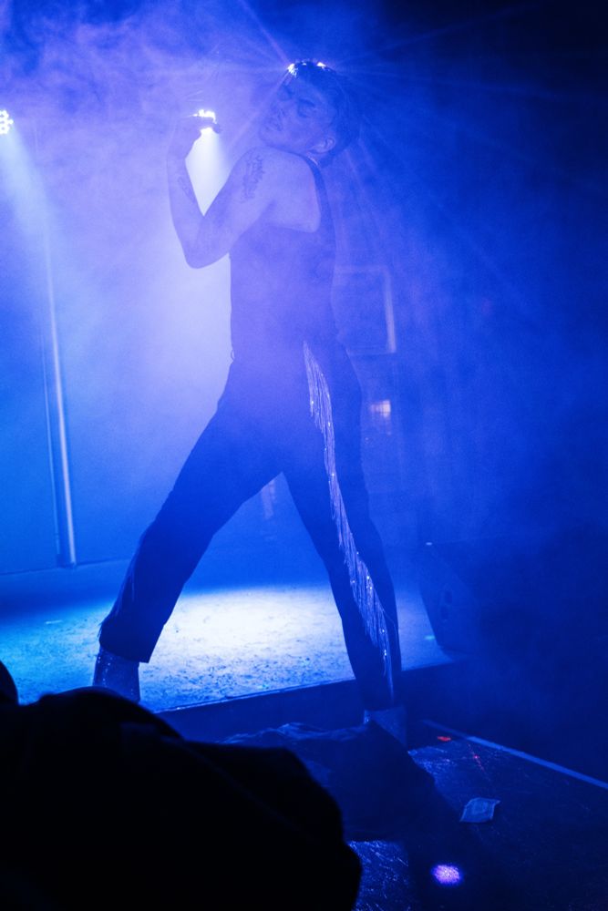 A blue/purple-hued photo of Layne, performing as Pam Jobb. They are standing on a catwalk with a stage behind and lights illuminating like a halo behind their head. Layne’s body is turned left while their head faces the audience, tilted up with their eyes closed. 
