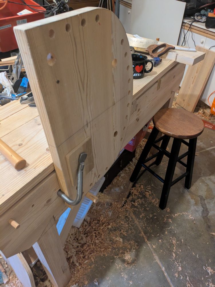 A chair seat is held awkwardly vertical to a workbench using a metal holdfast. 