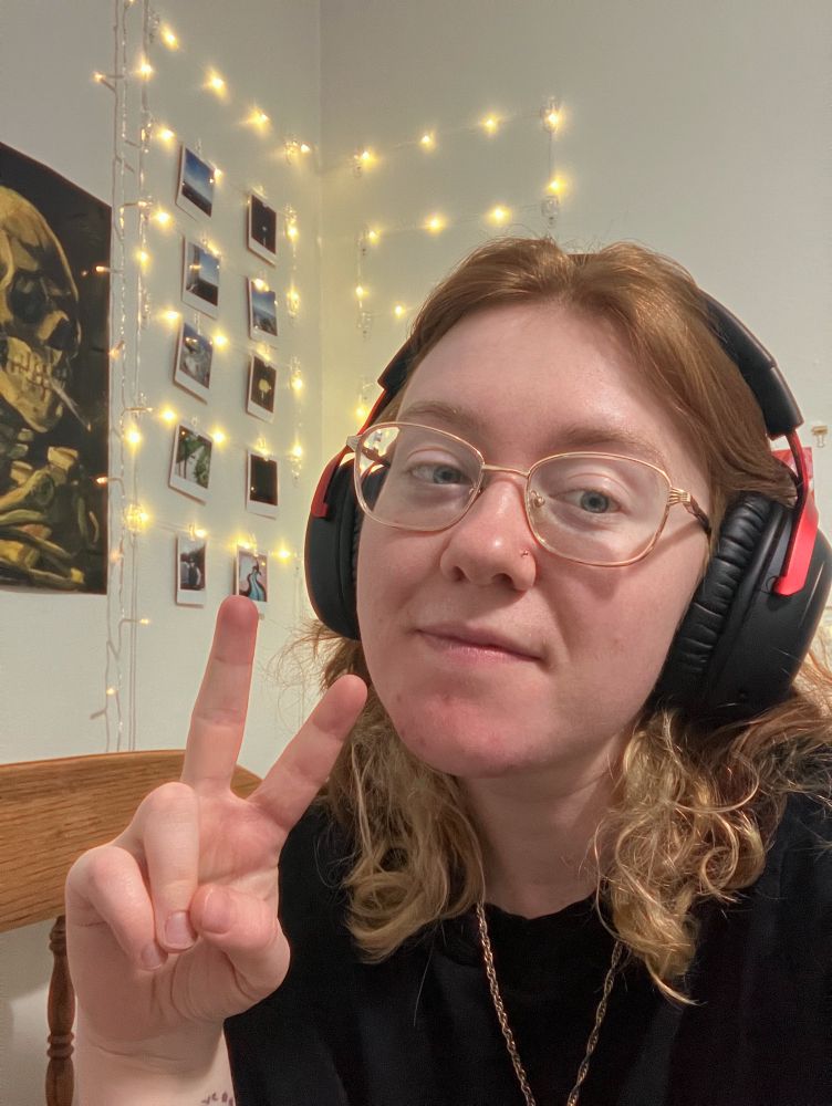 A selfie of me doing the peace sign, in front of my string light photo wall.