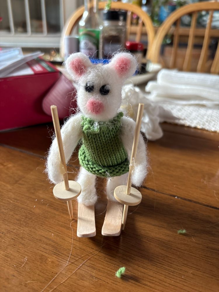 A white needlefelted mouse in a green knit sweater on wood skiis 