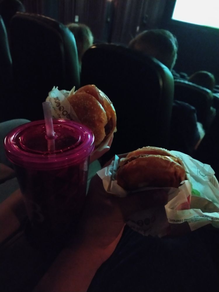 Foto colorida de dois sanduíches e um copo de refrigerante. Duas mãos seguram os sanduíches em volta de um copo grande de canudo e plástico rosa. O fundo da imagem é uma sala de cinema, onde é possível ver algumas cadeiras e parte da tela 
