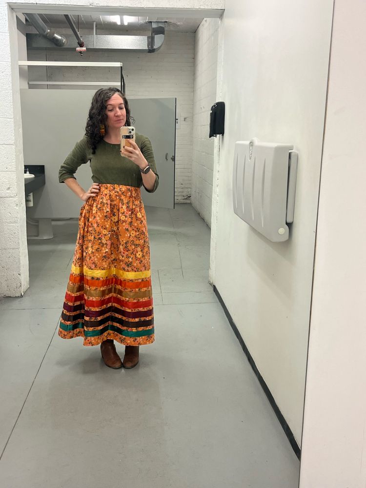 A brunette woman with curly hair takes selfie in full length bathroom mirror. She is wearing a yellow ribbon skirt and green top 
