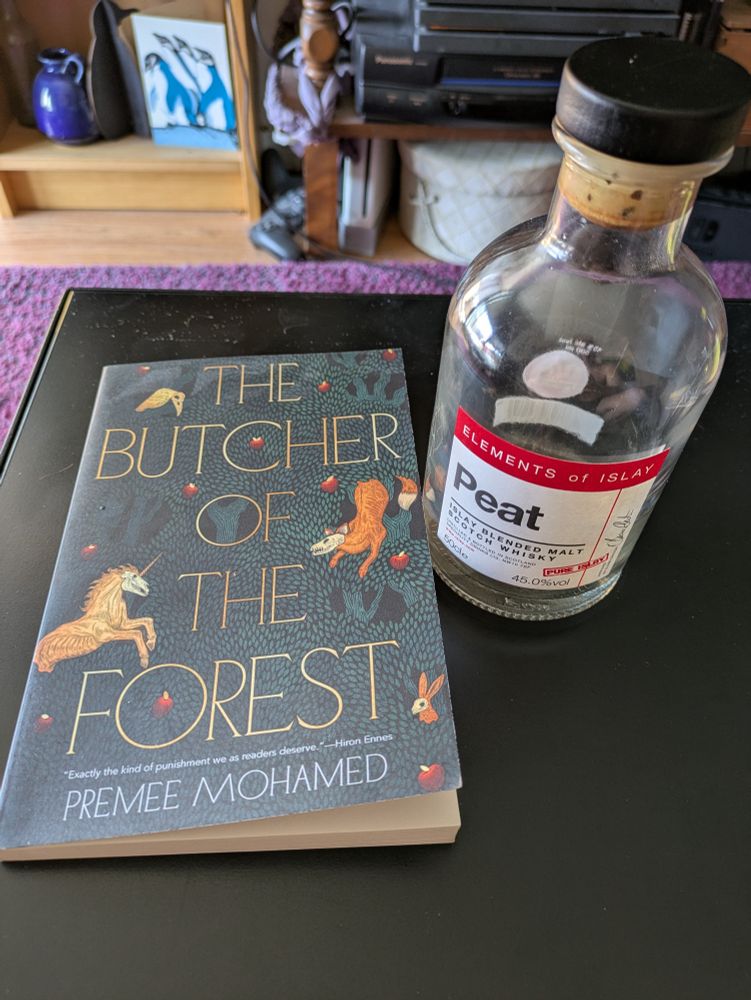 An (almost) empty bottle of blended scotch whisky with a period table of elements label reading "Peat" next to a copy of "The Butcher Of The Forest" by Premee Mohamed.