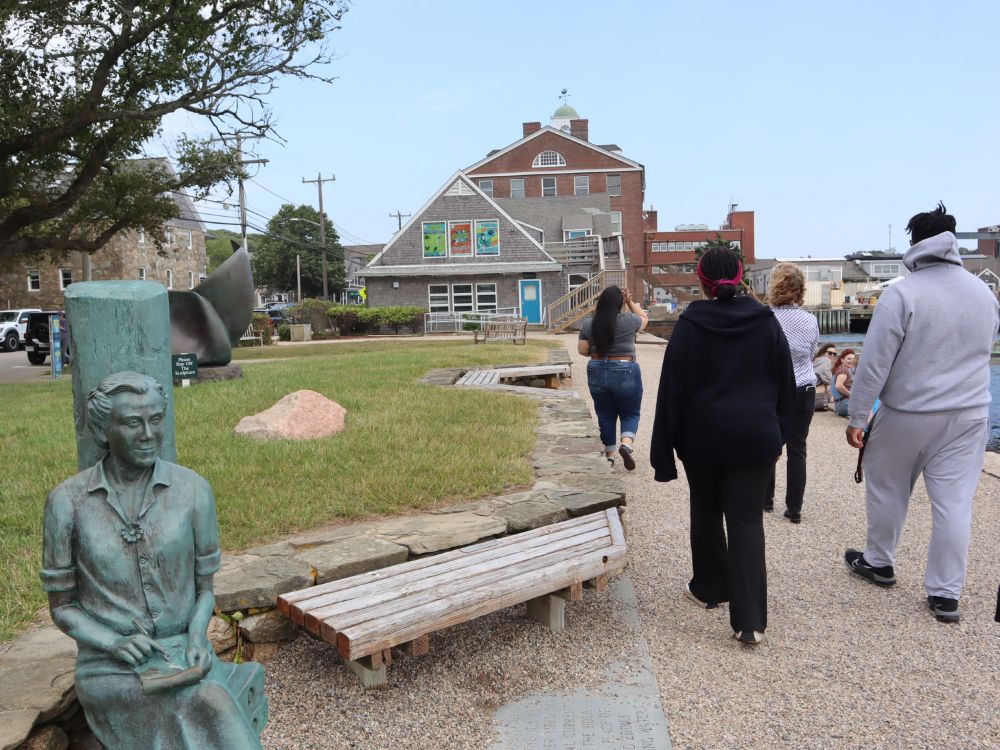 A group of people walk along a path on the waterfront. A statue of a seated woman is on their left.