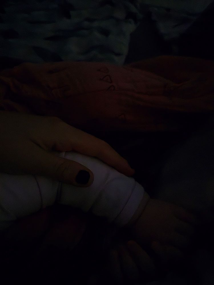 Ein verschwommenes, dunkles Bild zeigt eine Hand, die sanft den Unterarm von einem Baby berührt. Die Beleuchtung ist schwach und wirft weiche Schatten auf die Szene.
