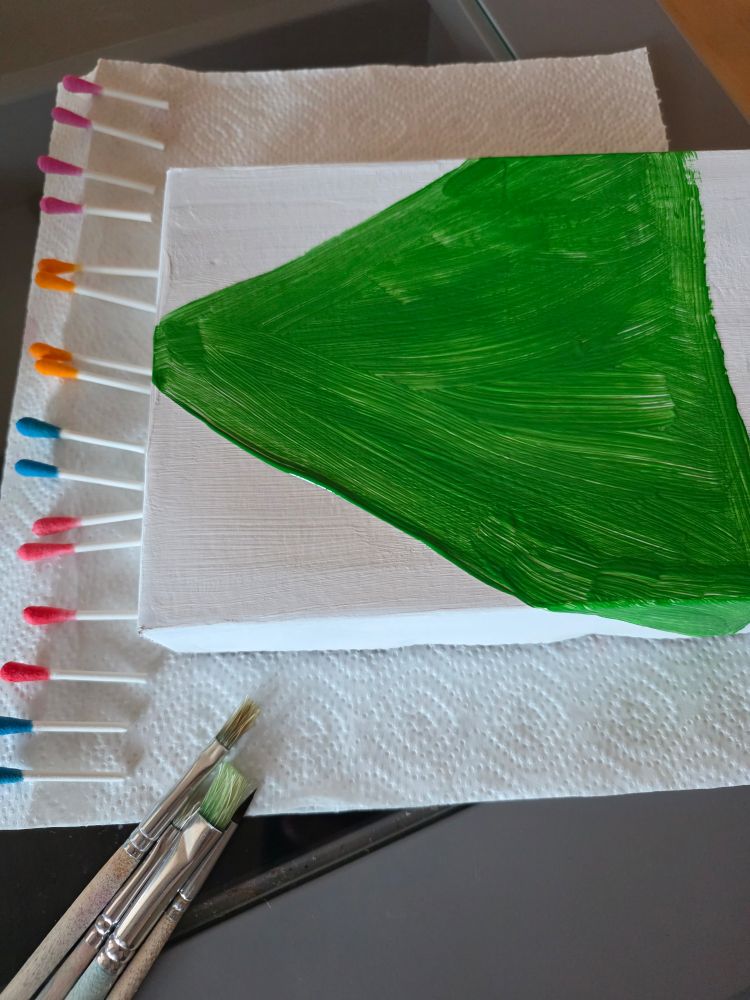 Ein weiß bemalter Schuhkartondeckel, auf dem ein leuchtend grünes Dreieck gemalt ist. Links daneben liegen Wattestäbchen, deren Enden verschiedenfarbig sind. Im Vordergrund sind einige Pinsel zu sehen. 