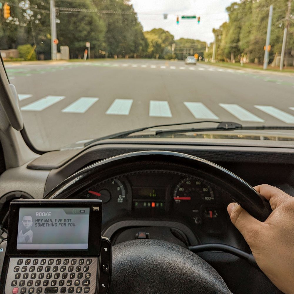 The interior of a vehicle at a stoplight, with a T-Mobile Sidekick keyboard phone visible in the corner. The Sidekick has a text message on the screen from Booke, a character in Midnight Club: Los Angeles, saying "Hey man, I've got something for you". This is the first text you receive from him, he being the final boss of the game.