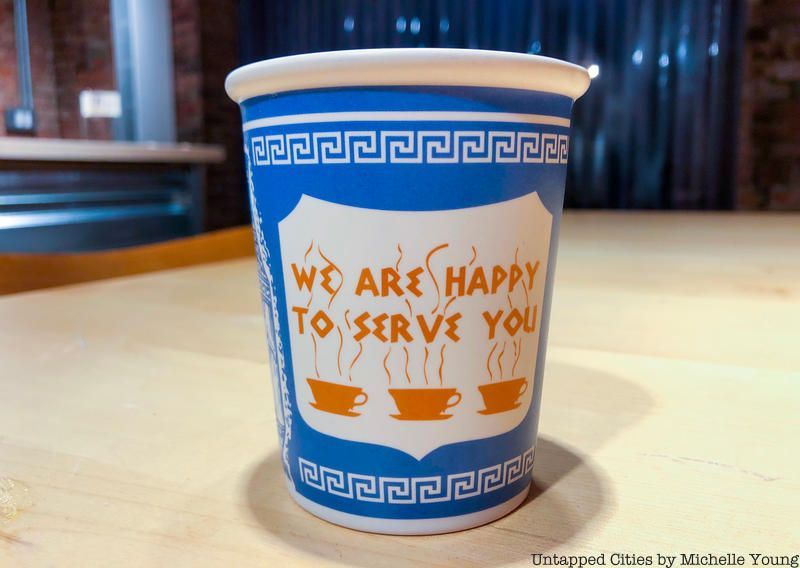 Greek diner paper coffee cup; a blue paper cup with text in a Greek-looking font that says, "We are happy to serve you" over top of images of three steaming coffee cups. There's a white on blue line motif around the top and bottom of the cup that's reminiscent of ancient Greek design and architecture.