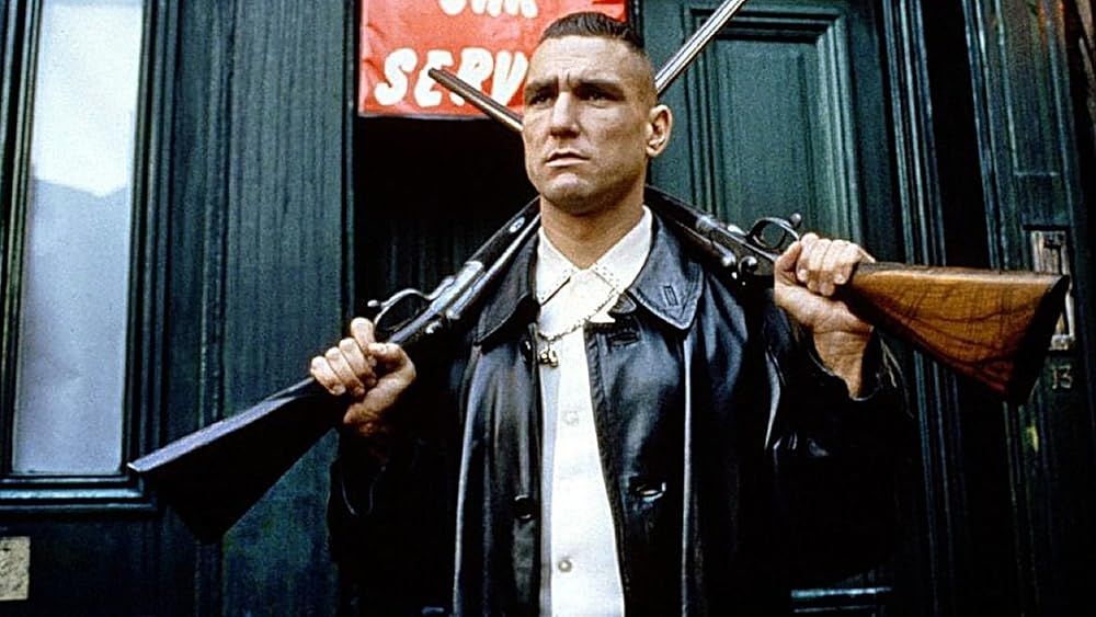 Vinnie Jones, a tall tough-looking guy, wearing a leather jacket and holding two rifles over his shoulders, probably in the movie Lock Stock and Two Smoking Barrels.