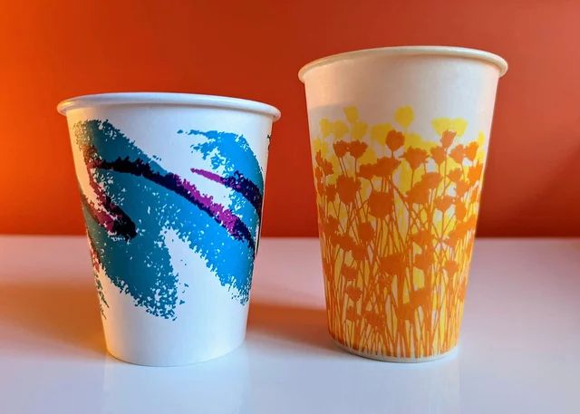 Two paper cups. On the left is the jazz cup; a white paper cup with a large blue and purple zigzag brushstroke pattern. On the right is a white paper cup with a design of lots of little yellow and orange flowers. 