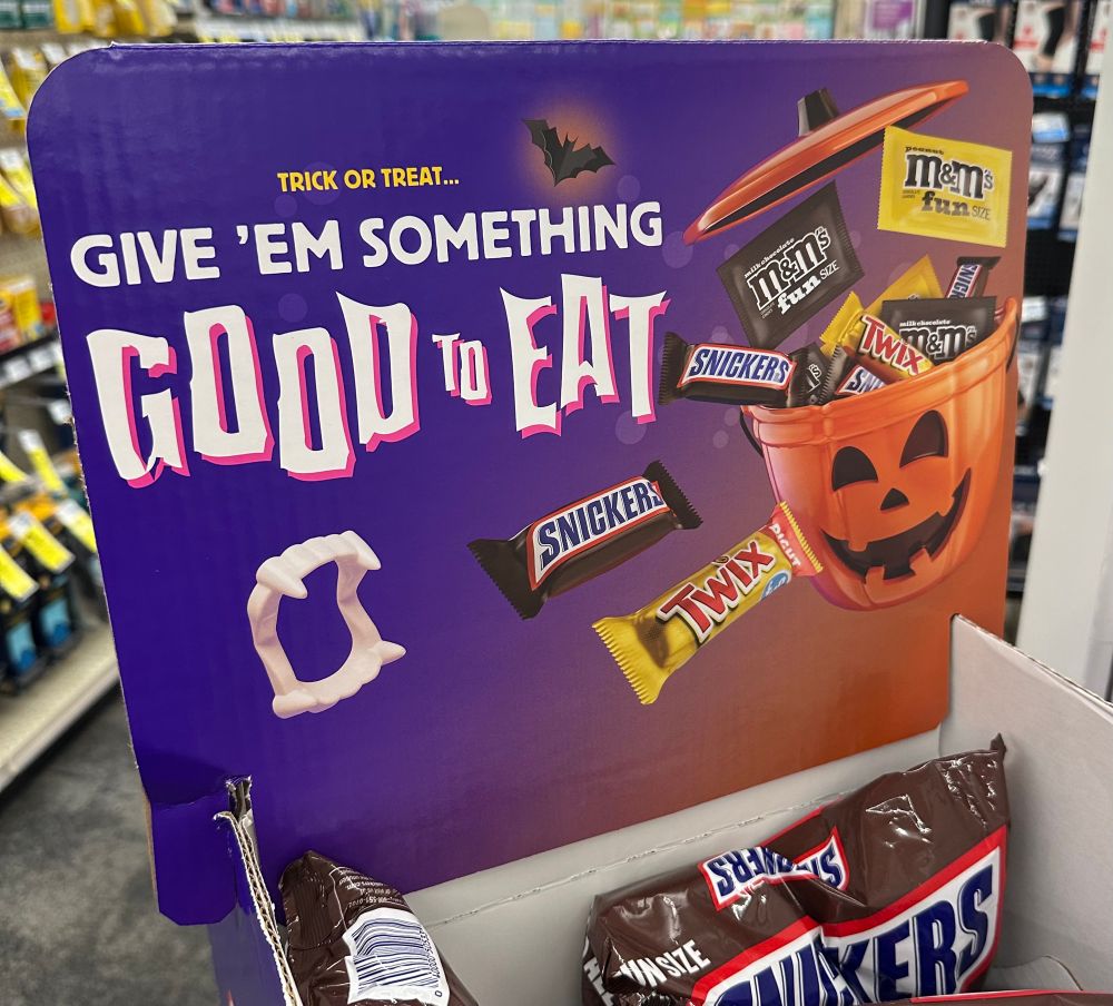 A display of Halloween candy in a drugstore. Details aren't important but it says, "Give 'em something good to eat," and there's a picture of a set of plastic vampire fangs and a plastic jack o' lantern bucket with candy popping out of it; Twix, Snickers, M&Ms, etc. Below the sign is a bag of Snickers. 
