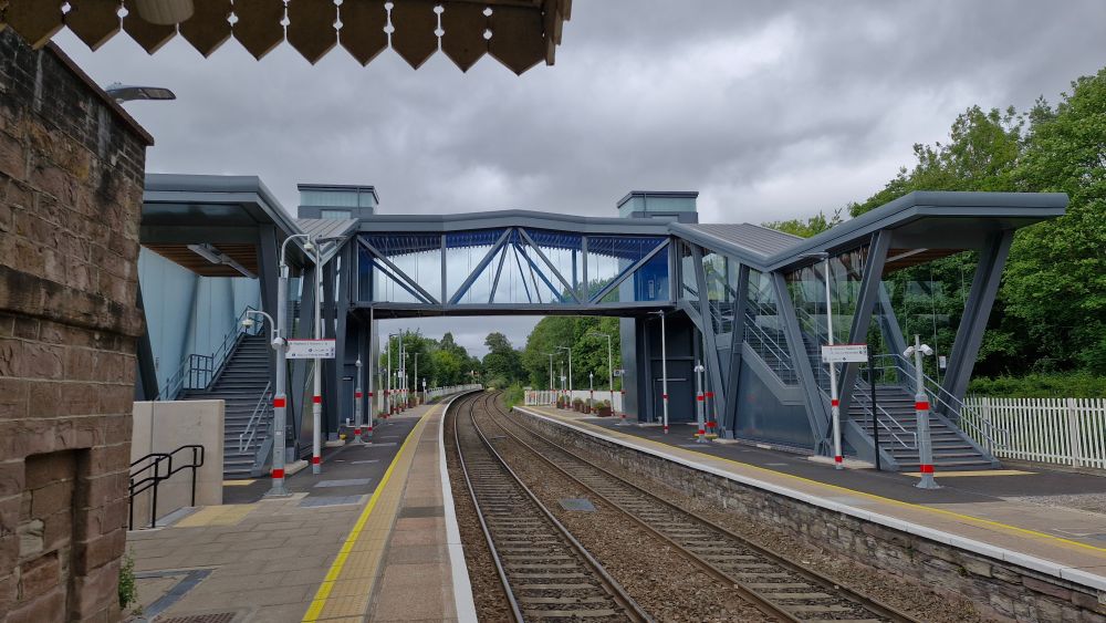 A modern footbridge across two railway tracks at a station, painted grey with glazed and roofed walkway with vertical and diagonal metal bracing visible. The lift towers behind have a clerestory on top of them.