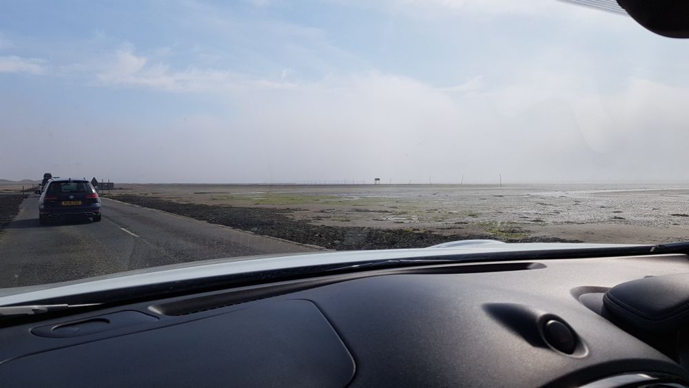 View across the dashboard of a car. A narrow strip of road has sand and water very close to it 