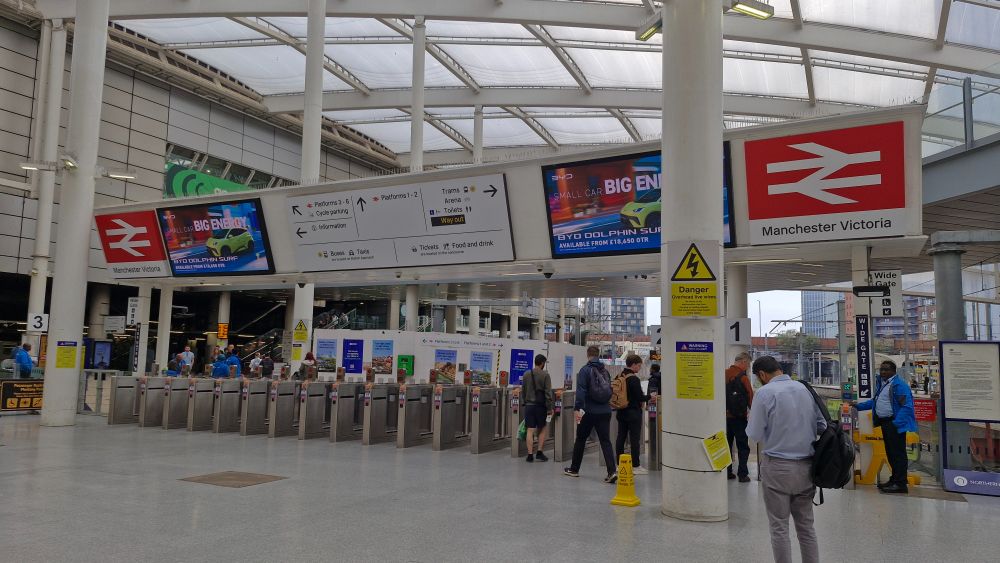 A railway station dateline with above it two very large double arrow logos next to LED advert screens with wayfinding signage in between.