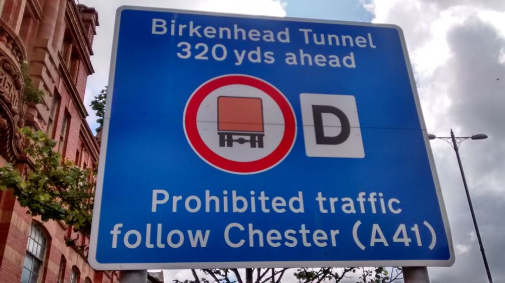 Sign Birkenhead Tunnel 320 yards ahead, red circle containing a stylised goods vehicle with a square orange body, letter D, Prohibited traffic follow Chester (A41).