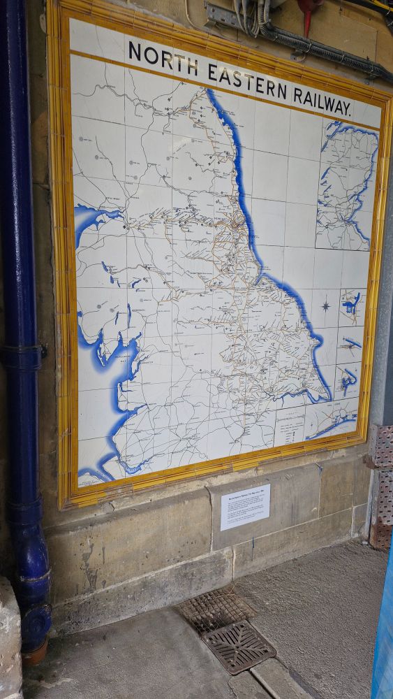 A map on tiles showing railway lines across the north of England, headed NORTH EASTERN RAILWAY. In a frame of orange-brown tiles, mounted on a stone wall. There is a small interpretation panel below the map.