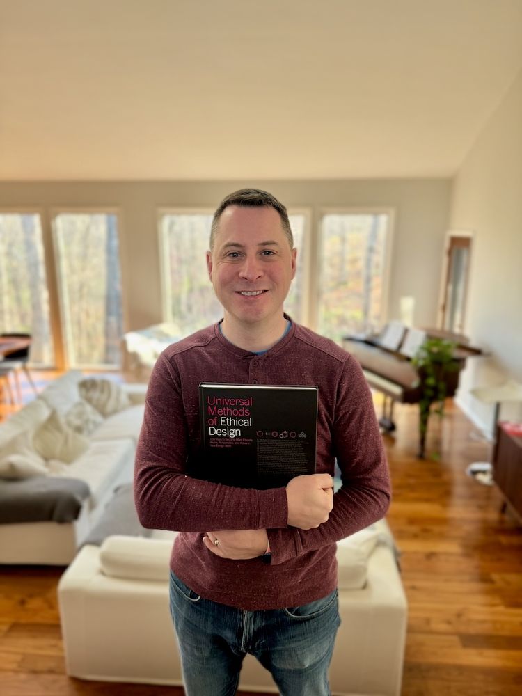 Colin is smiling, standing in a brightly lit living room holding a copy of Universal Methods of Ethical Design. 