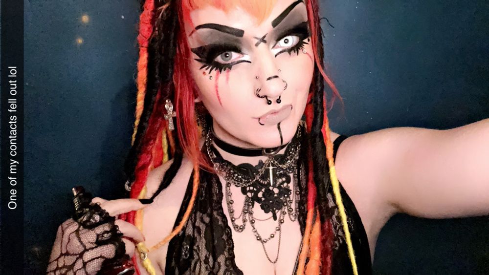 An alternative-styled girl takes a selfie. She has black, orange, red, and yellow hair. Multiple face piercings, and a black lace choker adorned with chains. She has one white contact in her left eye.