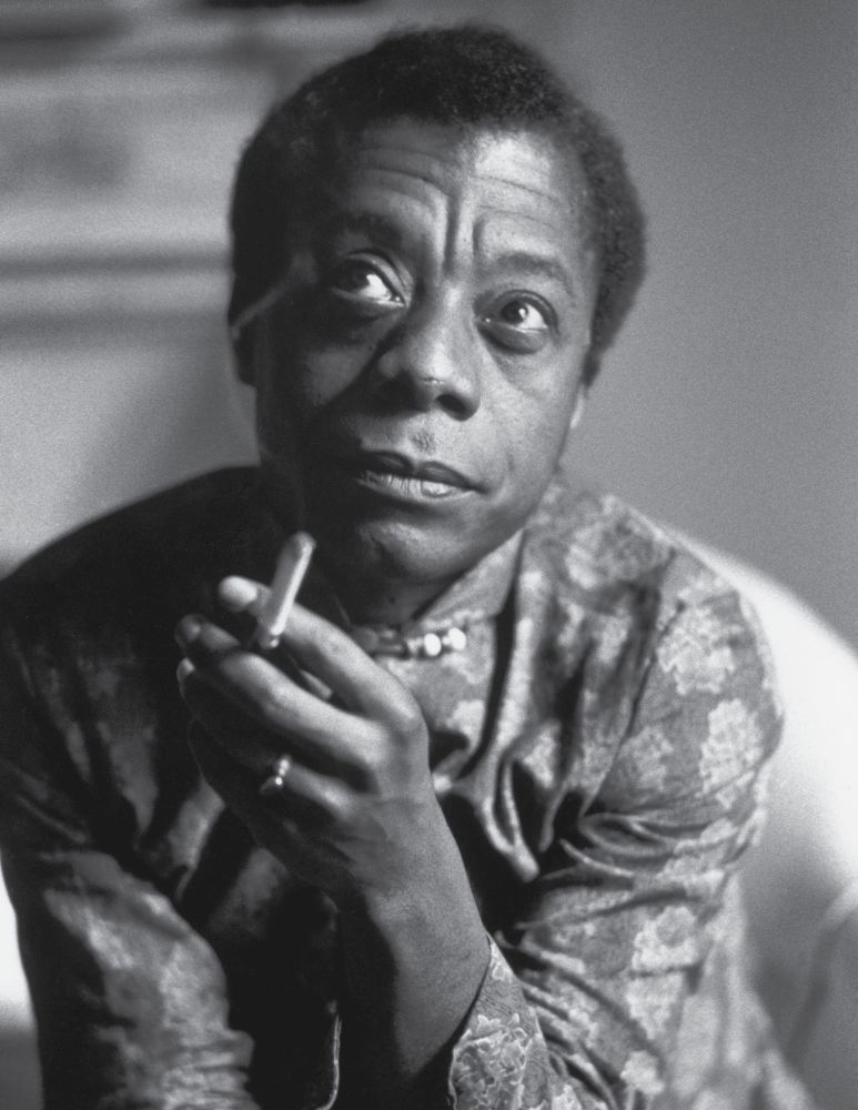 Black and white photo of James Baldwin facing the camera and looking up, holding a cigarette, wearing a patterned long sleeve shirt. 
