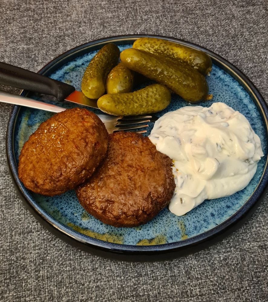 Kräuterquark, eingelegte Gurken und vegane Frikadellen auf einem blauen Teller.