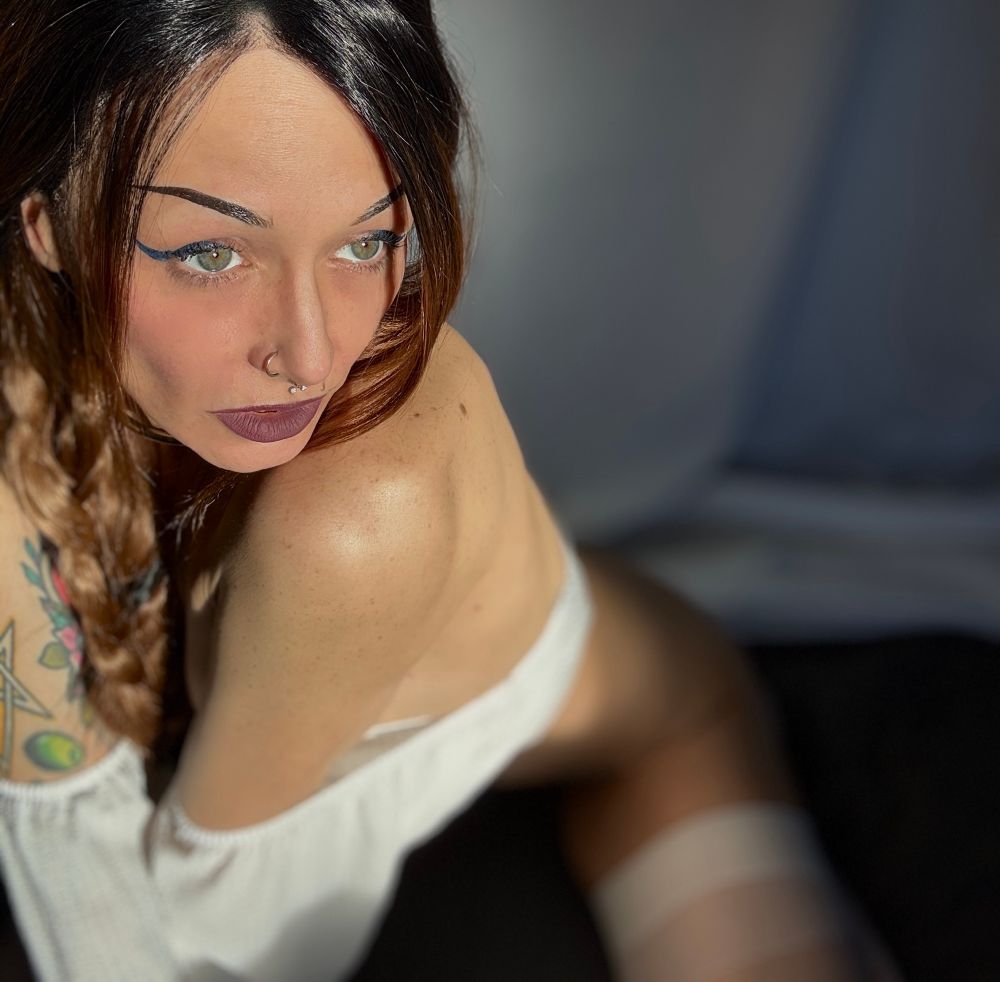 Fallen Angel Nat with straight brunette hair falling around her face, the bulk woven into a braid with black ribbons falling in front of her shoulder and down in-between her arms - they are extended with elbows locked propping her up as she leans forward with her face close to the lens. Her green eyes lines with blue taking focus. She wears only a white off the shoulder corset style top and sheer white seamed thigh highs- the curve of her bare waist, hips and ass seen in the background.