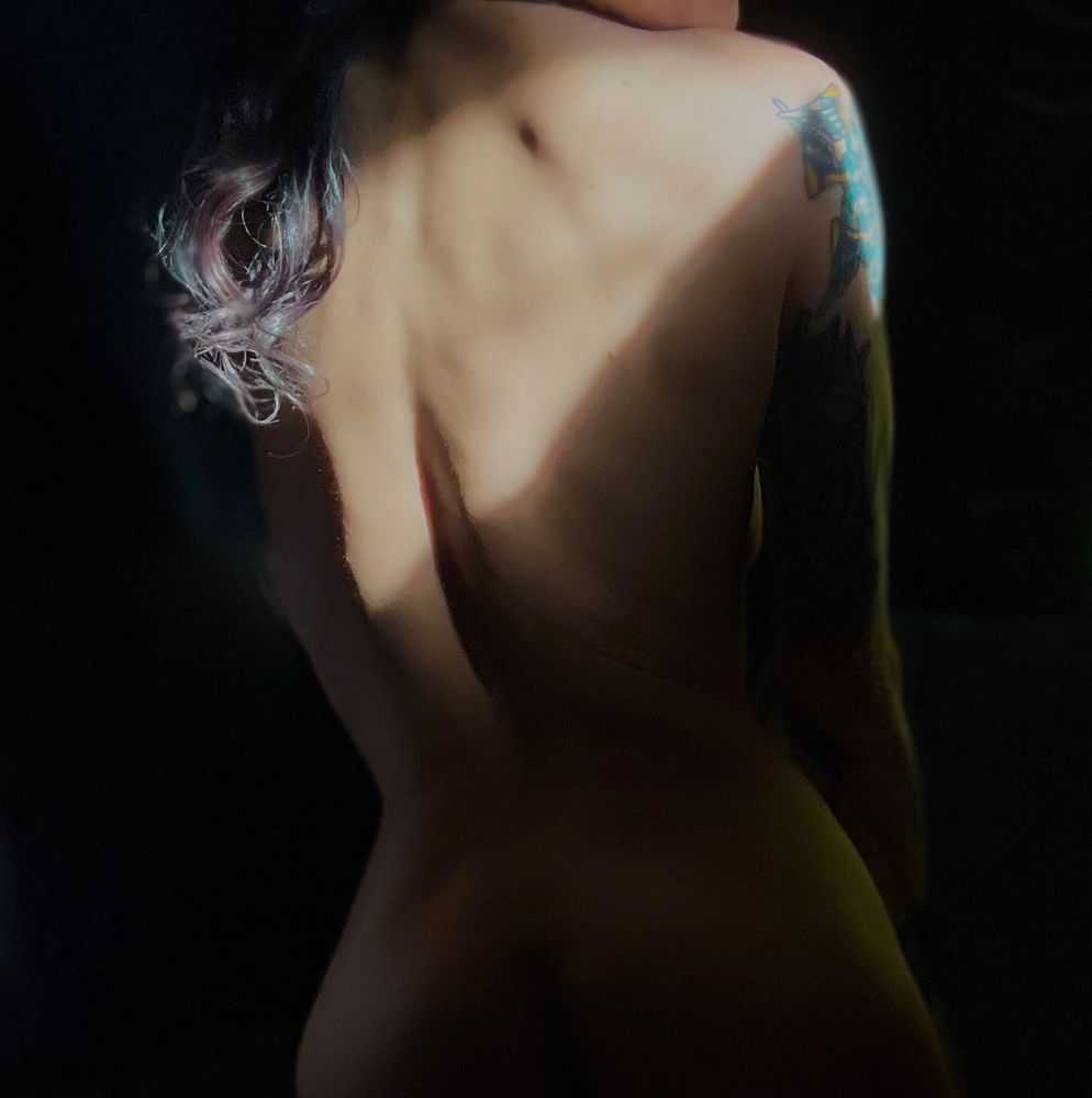 Her hourglass shape is front and center-Nat is seen turned away from the camera- in frame from the very top of her ass to the tops of her shoulders- she is heavily shadowed but for a slash of light across her back. Her red and blue curls fall over her shoulder frame left as her tattoo on the other shoulder catches a sliver of light.